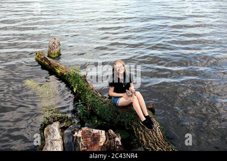 Junge blonde Frau mit blauen Haaren, Brille, Jeans-Shorts und einem schwarzen T-Shirt. Sitzt auf einem schwimmenden Baum in der Nähe des Sees mit einem drei-Monate-alt Stockfoto