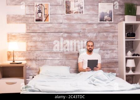 Fröhlicher Mann, der auf einem Tablet-Computer herumstöbert und nachts im Bett liegt. Stockfoto