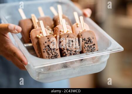 Verkäufer hält ein Tablett mit Schokoladeneis auf einem Stock im Laden, Nahaufnahme von Eis auf einer blauen Schürze Hintergrund Stockfoto