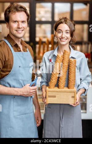 Porträt von zwei fröhlichen Mitarbeitern eines Cafés oder einer kleinen Konditorei, die mit einem Eiszapfen vor der Theke im Inneren stehen Stockfoto