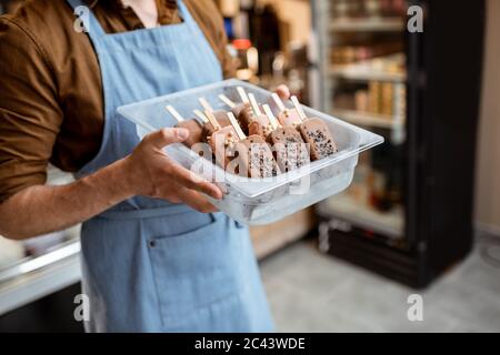 Verkäufer hält ein Tablett mit Schokoladeneis auf einem Stock im Laden, Nahaufnahme von Eis auf einer blauen Schürze Hintergrund Stockfoto