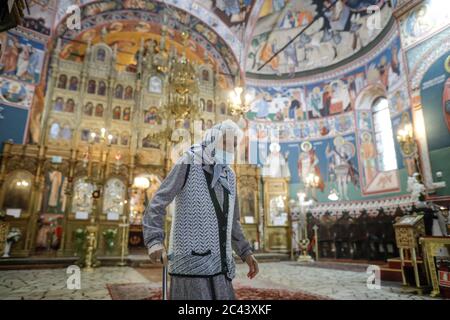 Snagov, Rumänien - 21. Juni 2020: Ältere Frau mit Covid-19 Maske betet vor einer orthodoxen christlichen Kirche. Stockfoto