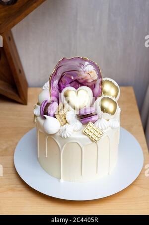 Moderner eleganter Kuchen mit geschmolzener weißer Schokolade, goldenem Herz und Schokoladenkugeln, Makronen, Baiser und Karamell. Stockfoto