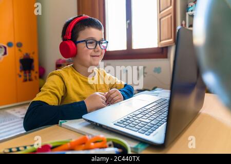 Lächelnder männlicher Student, der während der Heimsschooling über Kopfhörer hört und dabei auf den Laptop schaut Stockfoto