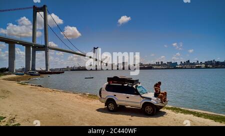 Mosambik, Katembe, Erwachsener Paar sitzt auf der Kapuze von 4x4 Auto bewundern Blick auf Maputo Bay mit Stadt und Maputo-Katembe Brücke im Hintergrund Stockfoto