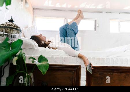 Lächelnde Frau, die auf dem Bett liegt und Musik mit Kopfhörern und Handy hört Stockfoto