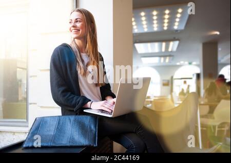 Glückliche Frau, die durch das Fenster schaute, während sie im Café einen Laptop benutzte Stockfoto