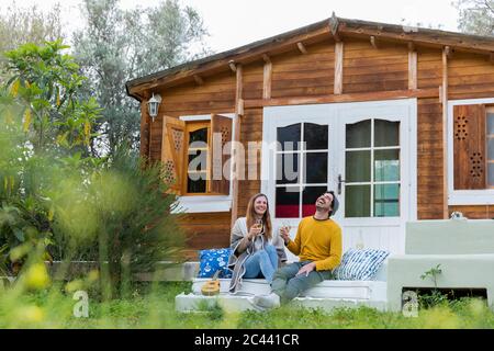 Fröhliches Paar, das Weingläser in der Hand hält und lacht, während es auf den Stufen der Blockhütte sitzt Stockfoto