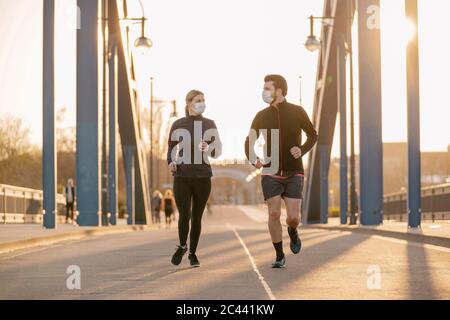 Junges Paar mit Gesichtsmasken läuft auf Brücke in der Stadt während des Sonnenuntergangs Stockfoto