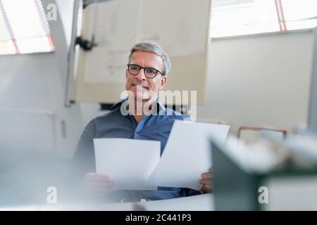 Lächelnd reifen männlichen Unternehmer sitzen mit Dokument, während am Schreibtisch im Büro zu betrachten Stockfoto