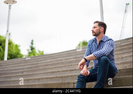 Blick aus der Nähe auf einen nachdenklichen Mann, der auf einer Amphitheatre in der Stadt gegen den Himmel sitzt Stockfoto