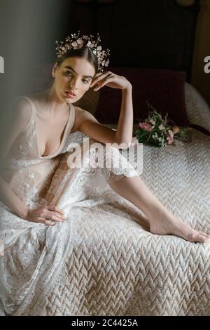 Junge Frau im Hochzeitskleid sitzt auf dem Bett und schaut seitwärts Stockfoto