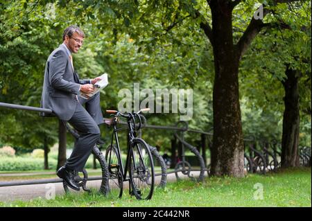 Geschäftsmann liest Zeitung, während er auf dem Geländer im öffentlichen Park sitzt Stockfoto