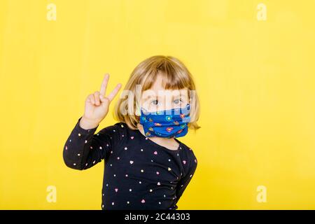 Portrait von niedlichen blonde Mädchen trägt Gesichtsmaske und gestikulierenden Frieden Zeichen vor gelbem Hintergrund Stockfoto