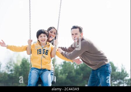 Fröhlicher Mann, der Mutter und Sohn auf Schaukel im Park gegen den klaren Himmel schiebt Stockfoto