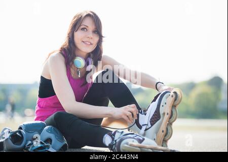 Lächelnde, selbstbewusste junge Frau, die Inline-Skates trägt, während sie im Park gegen den klaren Himmel sitzt Stockfoto