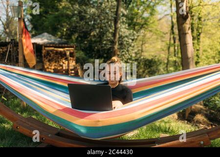 Junge Frau, die auf Laptop studiert, während sie in der Hängematte gegen Bäume sitzt Stockfoto