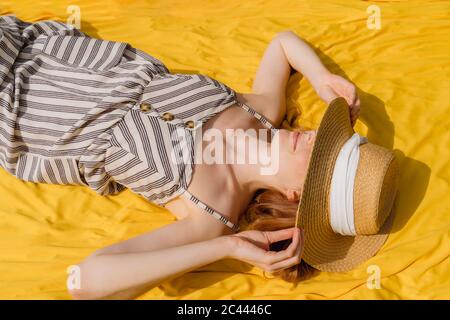 Junge Frau in Sundress hält Weidenhut, während sie auf gelbem Textil während sonnigen Tages liegen Stockfoto