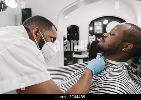 Friseur trägt chirurgische Maske und Handschuhe schneiden Bart des Kunden Stockfoto