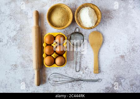 Nudelholz, Drahtquirl, Sieb, Hühnereier, Löffel und Schüsseln mit Mehl und braunem Zucker Stockfoto