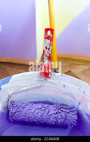 Walzenbürste in weißem Kunststoffschaft mit violetter Farbe, Nahaufnahme, im leeren Wohnzimmer, vertikales Bild Stockfoto