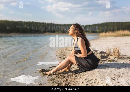 Junge Frau am Seeufer sitzend und Sonnenlicht genießend Stockfoto