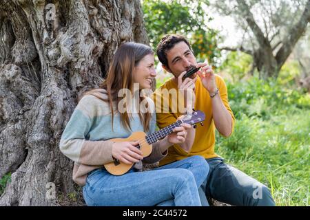 Glückliches Paar spielt Musikinstrumente, während sie am Baumstamm sitzt Stockfoto