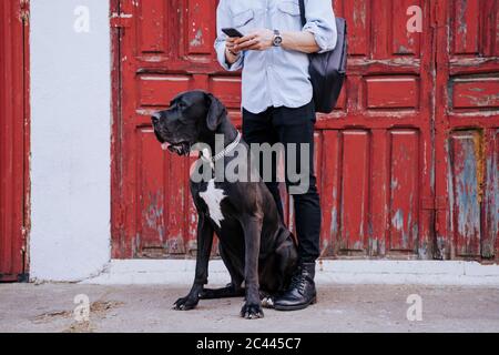 Crop-Ansicht von jungen Mann mit Handy wartet mit seinem Hund vor einer alten roten Holztür Stockfoto