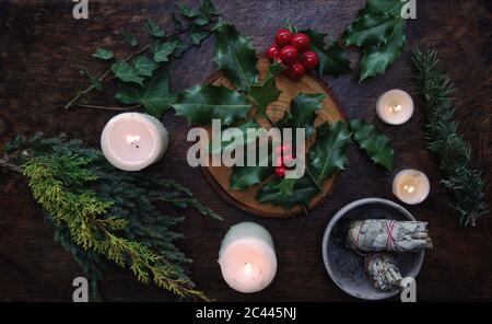 Yule Winter Sonnenwende (Weihnachten) thematische Wohnung lag mit Zweig der Stechpalme auf einem dunklen Holztisch. Salbei-Wischstöcke, weiß beleuchtete brennende Kerzen Stockfoto