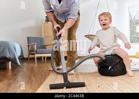 Niedriger Abschnitt des Mannes Reinigung Teppich, während nette Baby Junge sitzt auf Staubsauger im Wohnzimmer zu Hause Stockfoto