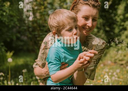 Glückliche Frau hält Sohn weht Löwenzahn Samen im Garten an sonnigen Tag Stockfoto