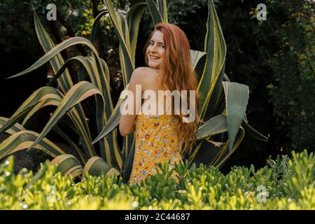 Lächelnde Frau mit langen braunen Haaren, die inmitten grüner Pflanzen im Garten steht Stockfoto