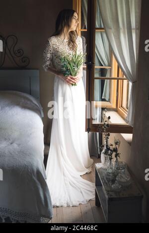 Junge Frau in eleganten Brautkleid hält Strauß Blick aus dem Fenster Stockfoto