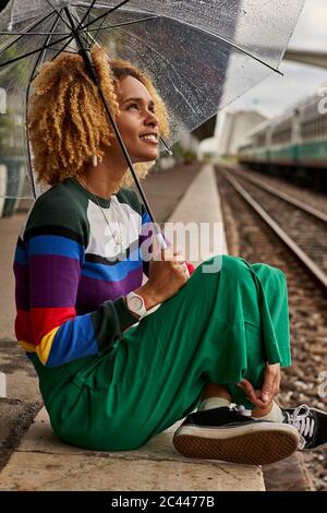 Volle Länge der jungen Frau sitzt mit gekreuzten Beinen auf Bahnhofsplattform während Monsun Stockfoto