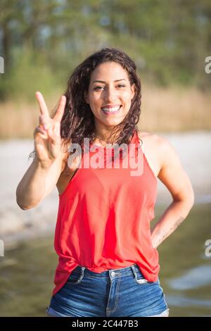 Porträt einer glücklichen jungen Frau am See mit Siegeszeichen Stockfoto