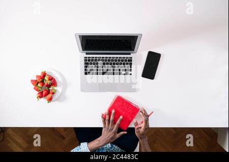 Ein junger Mann hält ein rotes Tagebuch am Laptop und ein Smartphone mit Erdbeeren am Schreibtisch Stockfoto
