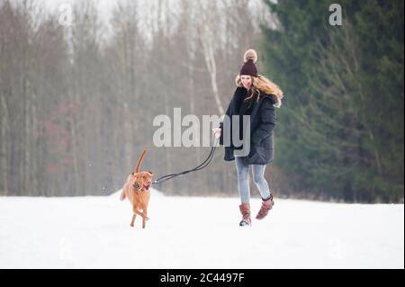 Glückliche junge Frau, die im Winter mit Hund auf Schnee gegen Bäume im Wald läuft Stockfoto