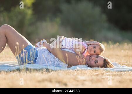 Portrait einer lächelnden Frau und Tochter, die während eines sonnigen Tages auf einer Picknickdecke auf der Wiese liegen Stockfoto