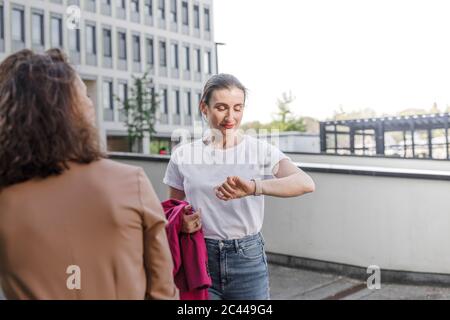 Geschäftsfrau beim Abschauen der Smartwatch, während sie mit Kollegen vor dem Bürogebäude steht Stockfoto