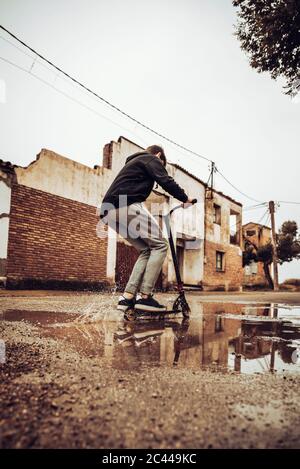 Teenager, der während der Regenzeit in der Pfütze auf der Straße mit einem Roller unterwegs ist Stockfoto