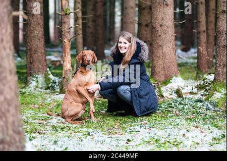 Lächelnde junge schöne Frau, die mit Hund gegen Bäume im Wald hocken Stockfoto