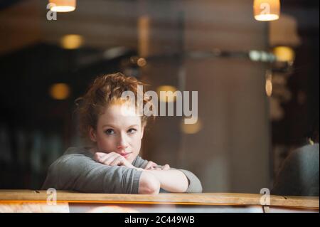 Nachdenkliche junge Frau, die sich durch das Glasfenster im Café auf den Tisch lehnt Stockfoto