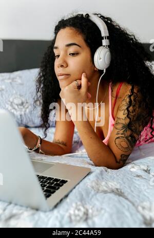 Schöne junge Frau im Bett zu Hause mit Kopfhörern und mit Laptop liegen Stockfoto