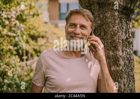 Lächelnder reifer Mann schaut weg, während er auf dem Handy gegen den Baum im Garten spricht Stockfoto