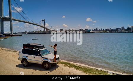 Mosambik, Katembe, Erwachsene Frau mit erhobenen Armen auf Stoßstange von 4x4 Auto bewundern Blick auf Maputo Bay mit Stadt und Maputo-Katembe Brücke im Hintergrund Stockfoto