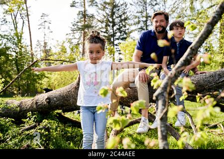 Nettes Mädchen mit ausgestreckten Armen stehen gegen Vater und Bruder auf gefallenen Baum im Wald sitzen Stockfoto