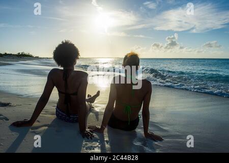 Ansicht der Rückseite des weiblichen Freunden am Meer sitzend auf Grace Bay Strand bei Sonnenuntergang, Providenciales, Turks- und Caicosinseln Stockfoto