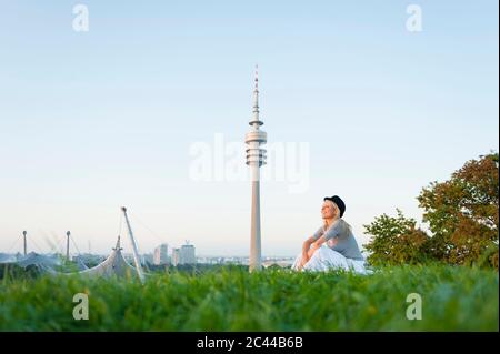 Nachdenkliche junge Frau auf Gras sitzend mit Olympic Tower im Hintergrund in München, Deutschland Stockfoto