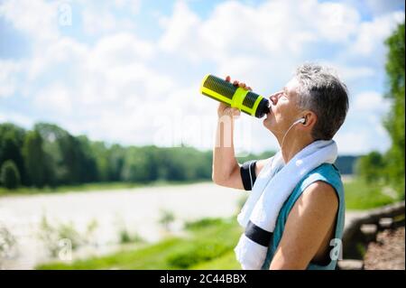Seitenansicht des durstigen aktiven älteren Mannes, der Wasser aus der Flasche am Flussufer trinkt, an einem sonnigen Tag Stockfoto