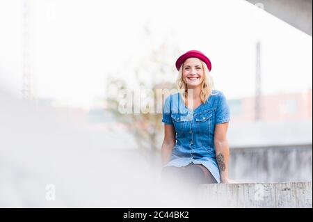 Porträt einer glücklichen jungen Frau in Jeanskleid an einer Wand sitzend Stockfoto